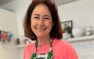 Kat smiling to camera, wearing a pink jumper and a green floral patterned kitchen apron