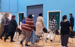 Rows of people stood behind chairs in a gym, lifting legs behind them, with instructor at the front