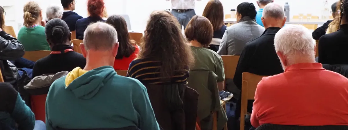 The MC stood at the far end of the room, looking at the camera over the heads of his seated audience 