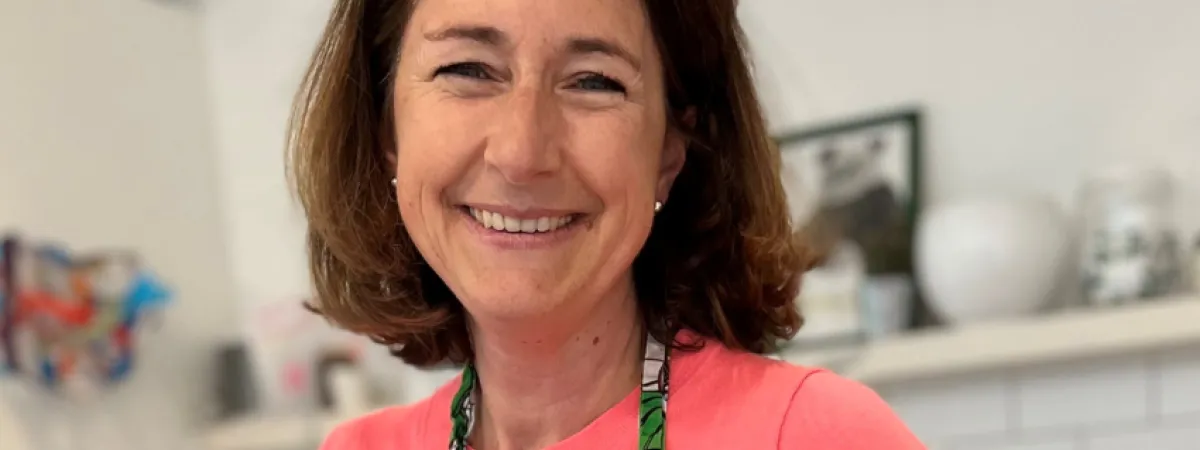 Kat smiling to camera, wearing a pink jumper and a green floral patterned kitchen apron