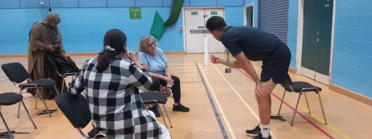 Two women sat down on chairs in the gym, with cups of tea, talking to a male fitness instructor