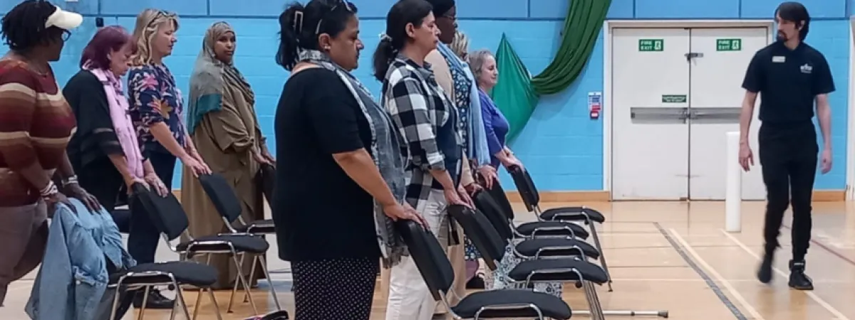 Rows of people, stood behind chairs, looking to the front of the gym at their instructor