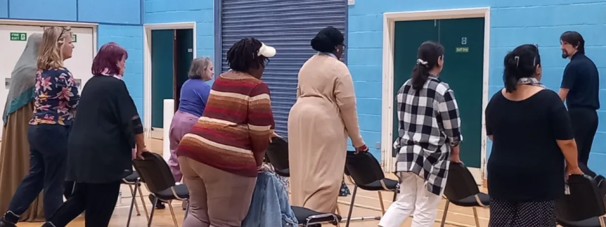 Rows of people stood behind chairs in a gym, lifting legs behind them, with instructor at the front