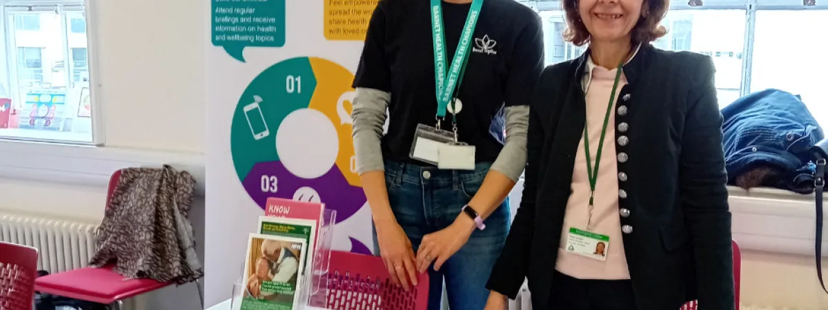 Two women smiling at a table with health leaflets. Sign reads Barnet Health Champions. 