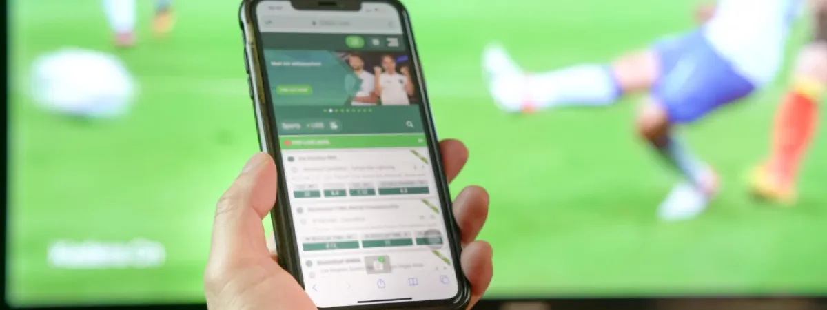 Someone holding a mobile phone, on which a betting app is open, with football on screen in the background.